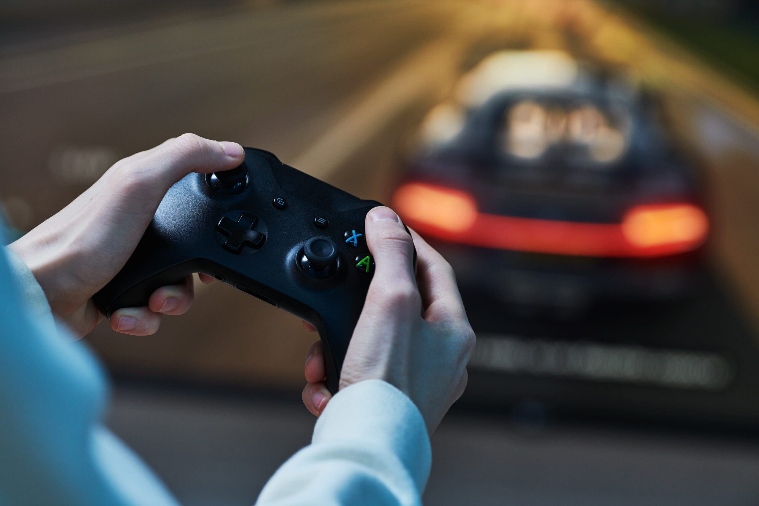 Close-up of hands holding a black gaming controller, with a blurred racing game on screen in the background.